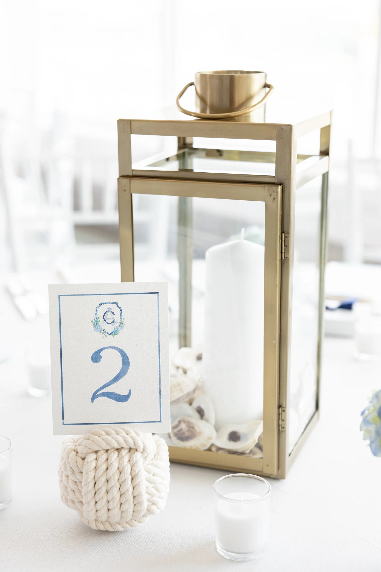 Gold lantern centerpiece with oyster shells and  a candle, handpainted blue table number card, and decorative rope table number holder.