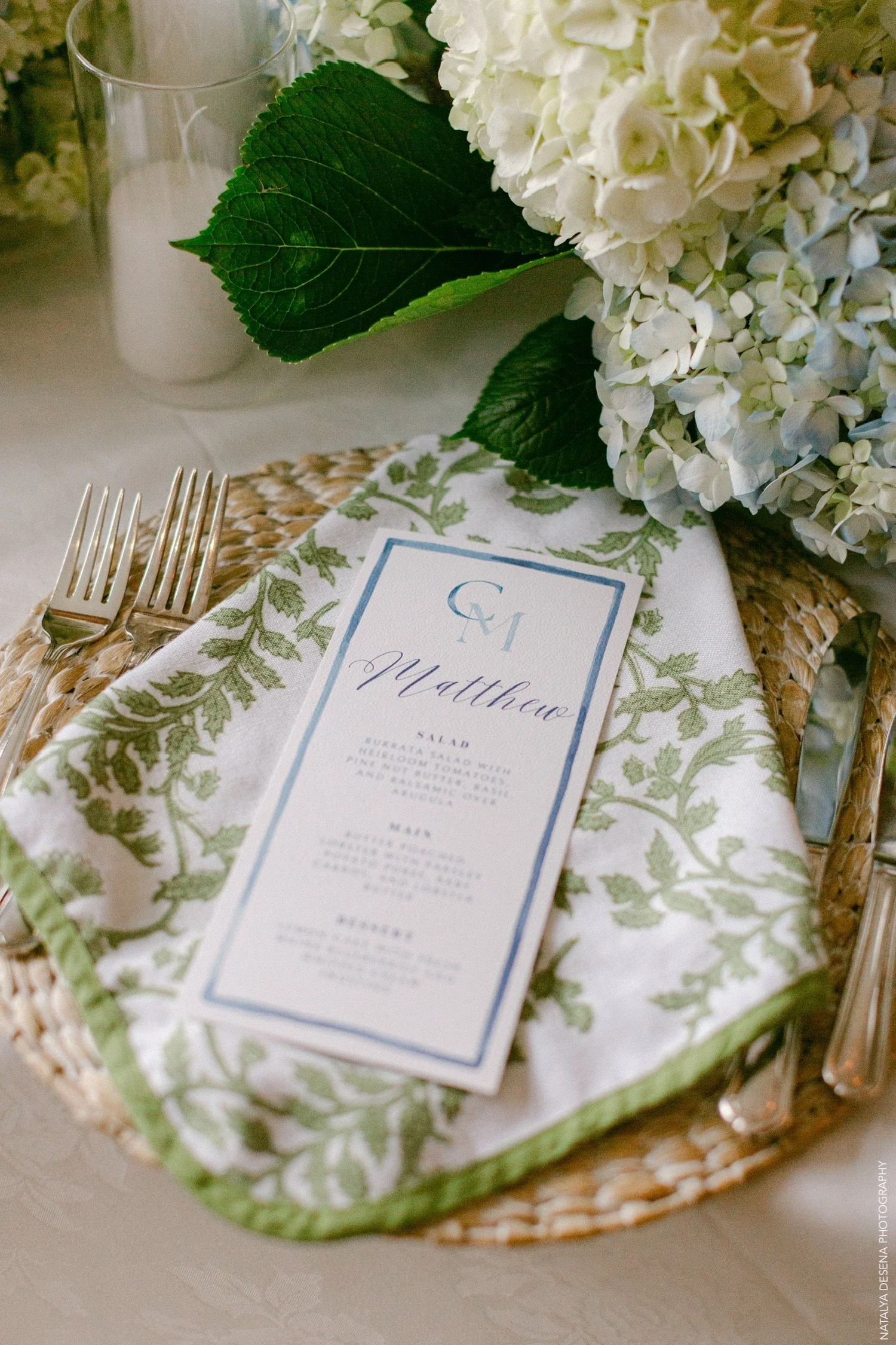 Table setting with floral arrangement, custom monogram menu card, and cutlery on a woven placemat.