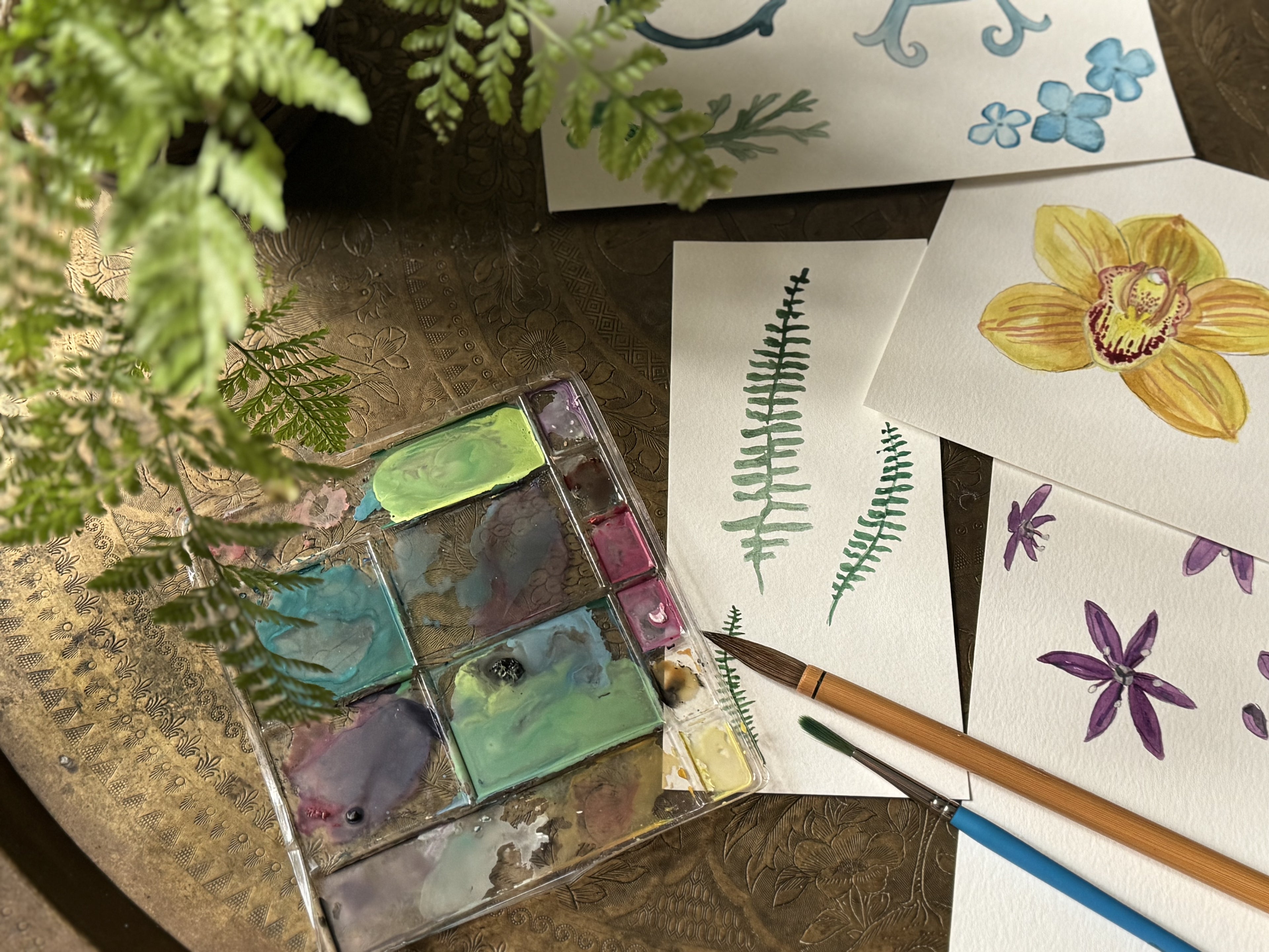 Art supplies including watercolor paints, brushes, and watercolor samples on a wooden surface.